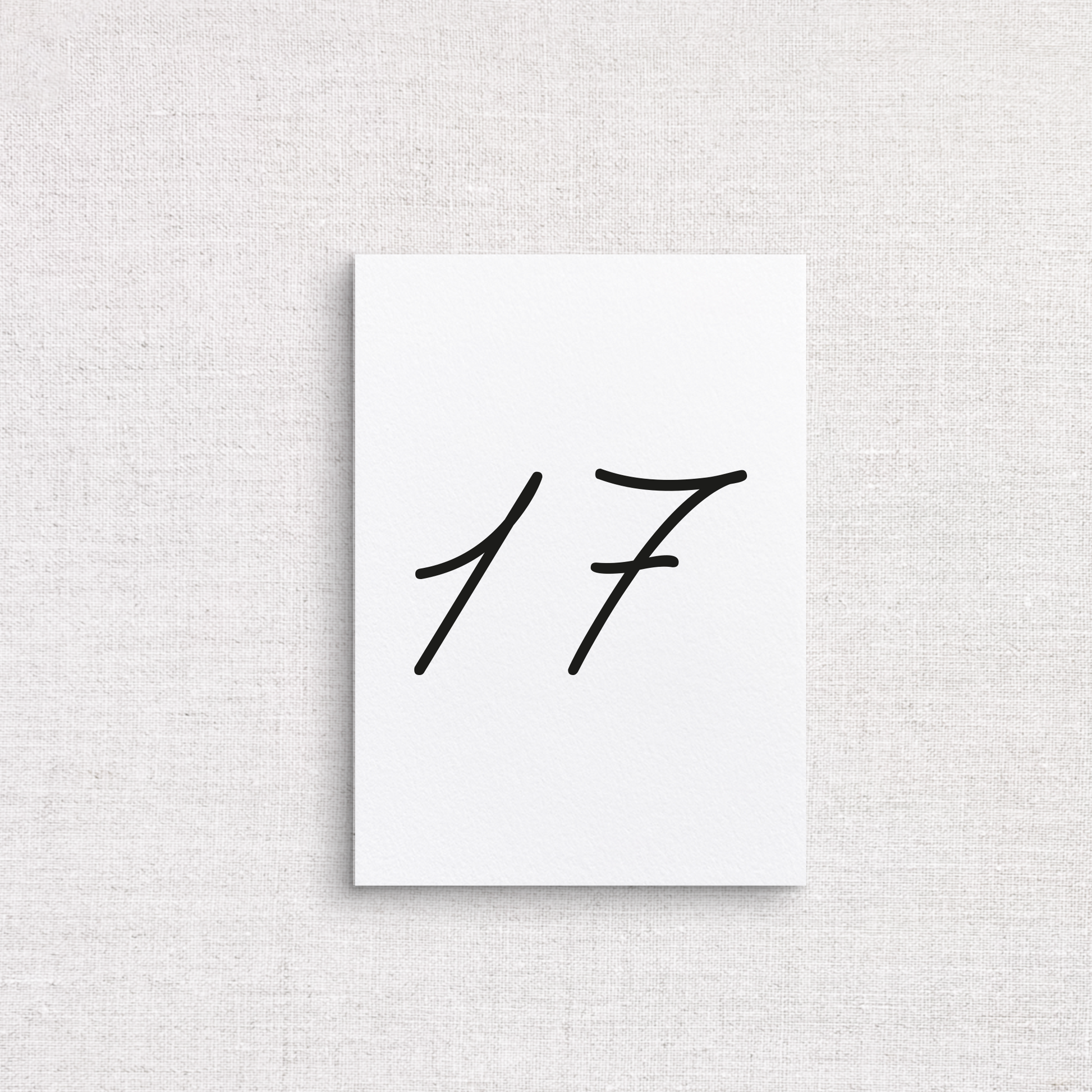 A minimalist white wedding table number featuring the black handwritten-style numbers '17', centered on a light beige textured fabric background.