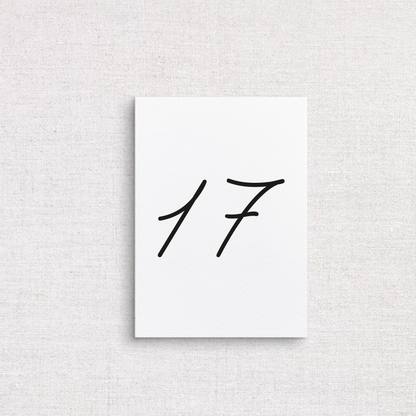 A minimalist white wedding table number featuring the black handwritten-style numbers '17', centered on a light beige textured fabric background.