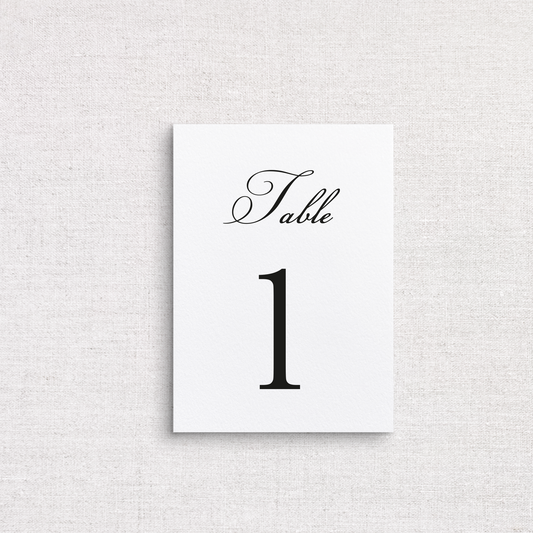 A vertical, white, traditional table number card reading 'Table 1' in black script and serif font, centered on a light beige textured fabric background.