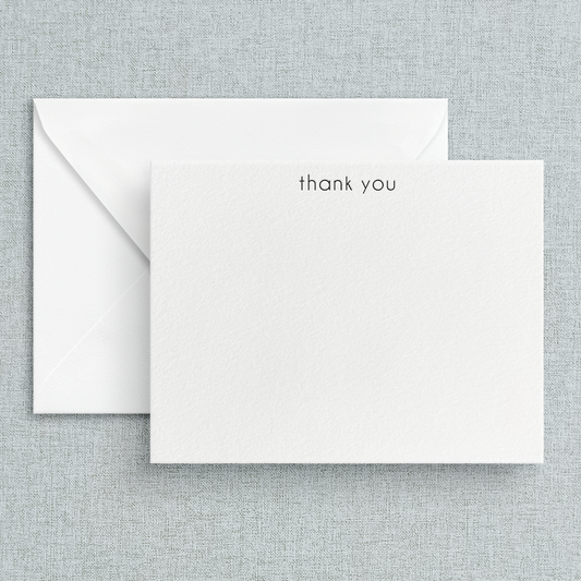 A simple letterpress printed 'thank you' note printed in lowercase on a white note card, accompanied by a white envelope, set against a textured background.