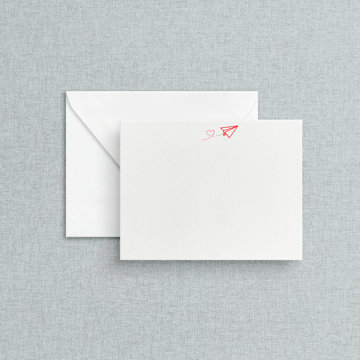 A letterpress stationery flat card with red airplane with a heart trail print on it accompanied by an envelope, placed against a textured grey background.