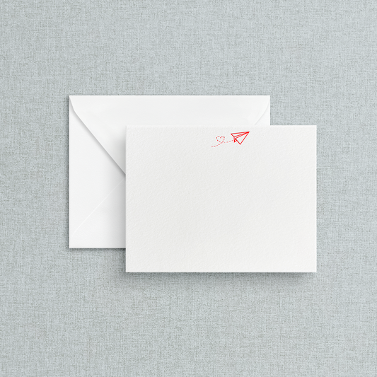 A letterpress stationery flat card with red airplane with a heart trail print on it accompanied by an envelope, placed against a textured grey background.