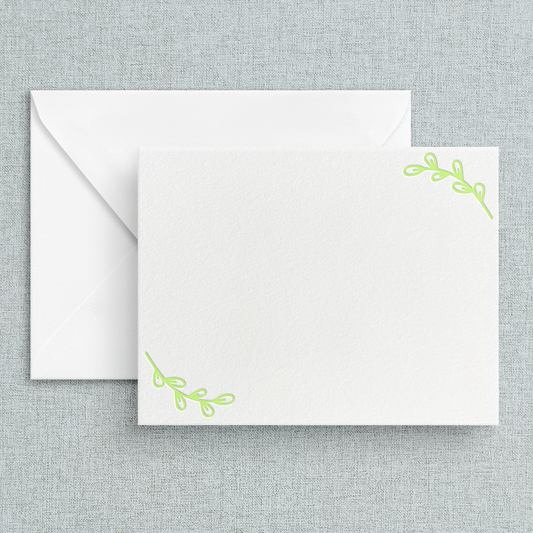 A set of white letterpress stationery cards with sage green leaf designs in the corners, accompanied by white envelopes, displayed against a textured background.